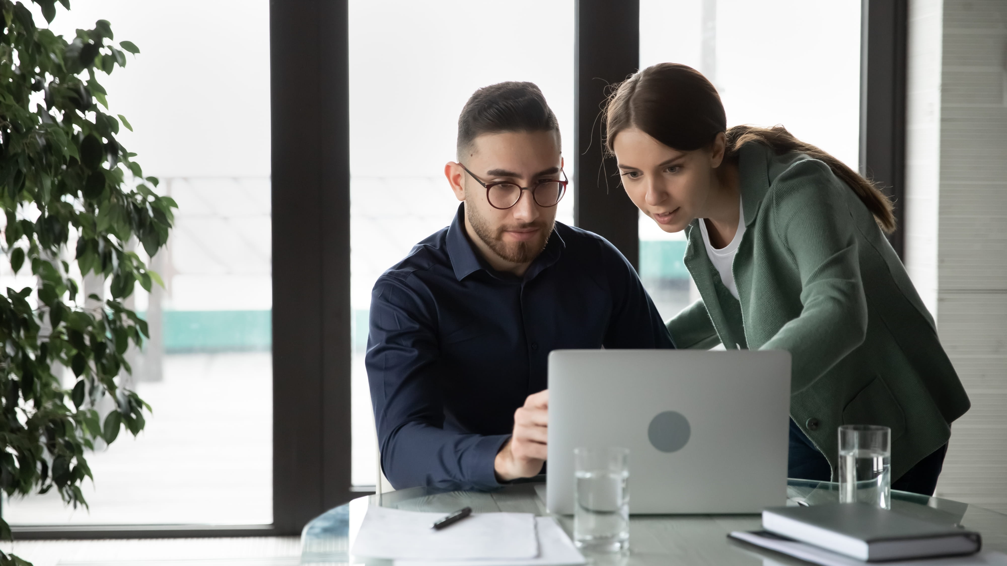 Man in blue shirt and woman in green hoodie consider how to choose a corporate caterer while looking at a laptop.